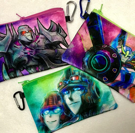 Sample of handmade velvety plush zippered mini bags. Photo shows iverillian's illustrations of Megatron from Transformers Prime, Strongarm from Robots in Disguise, and Lug & Anode from IDW Lost Light.